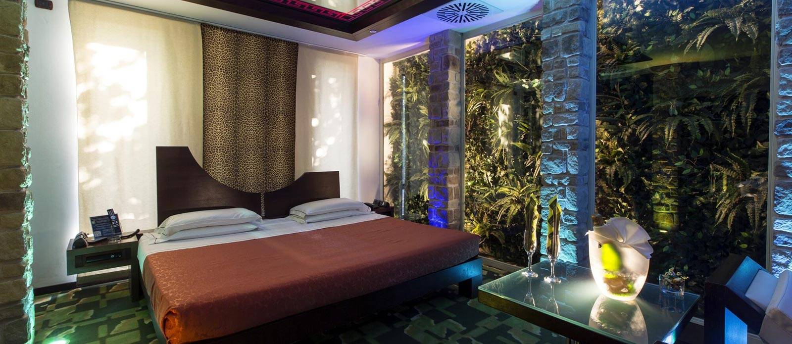 Motel K Wellness Suite Pietra del Sol vista sul letto matrimoniale con specchio a soffitto, bottiglia di champagne in secchiello del ghiaccio sul tavolo di vetro davanti al letto assieme a 2 bicchieri, camera a tema antica civiltà azteca 